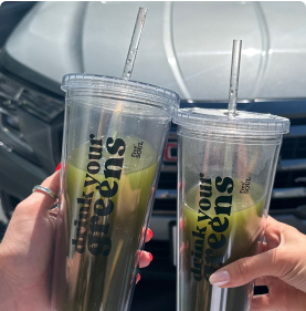 Two hands holding clear cups labeled 'drink your greens' with green juices inside.