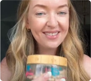 Woman smiling and holding a jar with a wooden lid, containing colorful objects inside.