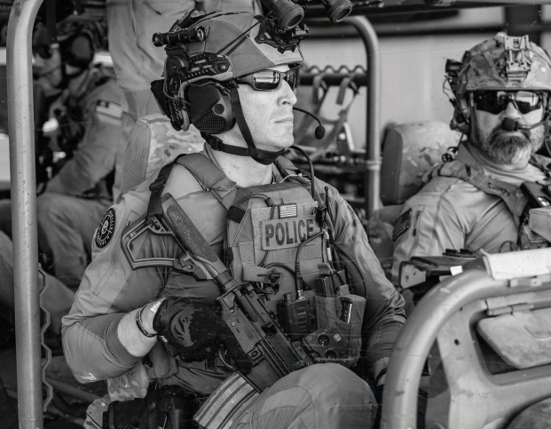 Police tactical unit members in military gear seated in a vehicle.