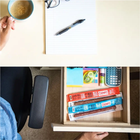 Desk setup with coffee, notepad, pen, open drawer with snacks and office supplies.