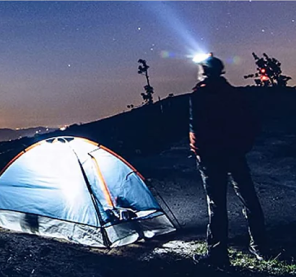 Person with headlamp beside illuminated tent under starry sky.