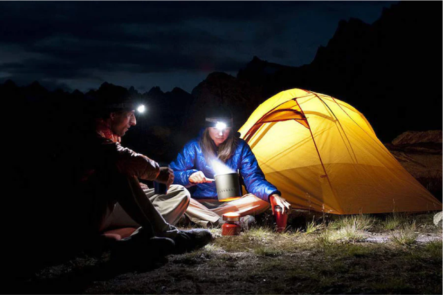 Two campers with headlamps cooking near a yellow tent at night.