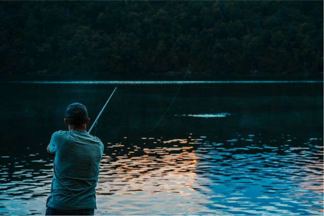 Person fishing by a calm lake at sunset.