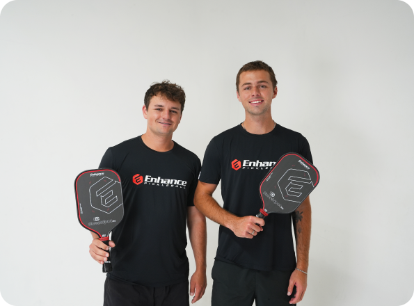 Two men smiling, holding pickleball paddles, wearing matching Enhance shirts, against a plain background.