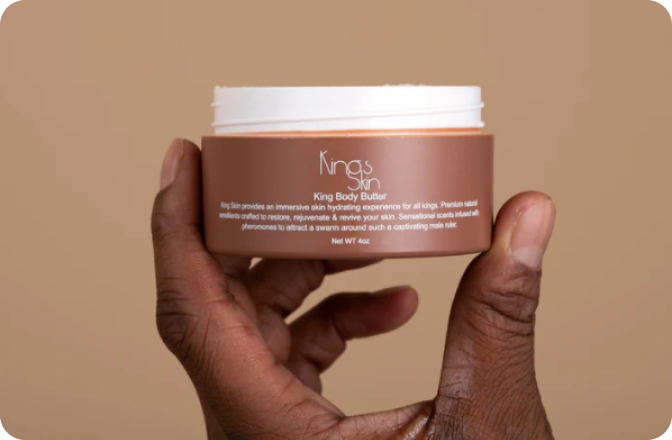 A hand holding a container of King Body Butter against a beige background.