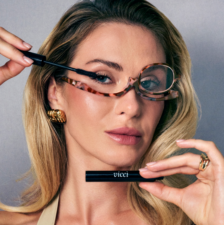 Woman applying mascara with glasses; holding a tube labeled 'vicci.'