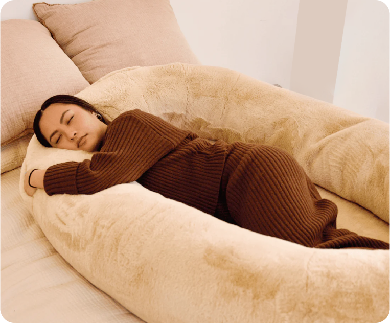 Woman sleeping comfortably in a beige body pillow on a bed.