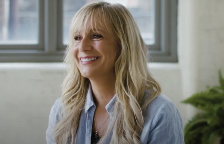 Smiling woman with long blonde hair in a light blue top, sitting indoors near a window.