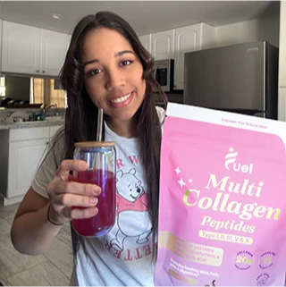 Woman in kitchen holding a drink and collagen peptide package, smiling at camera.