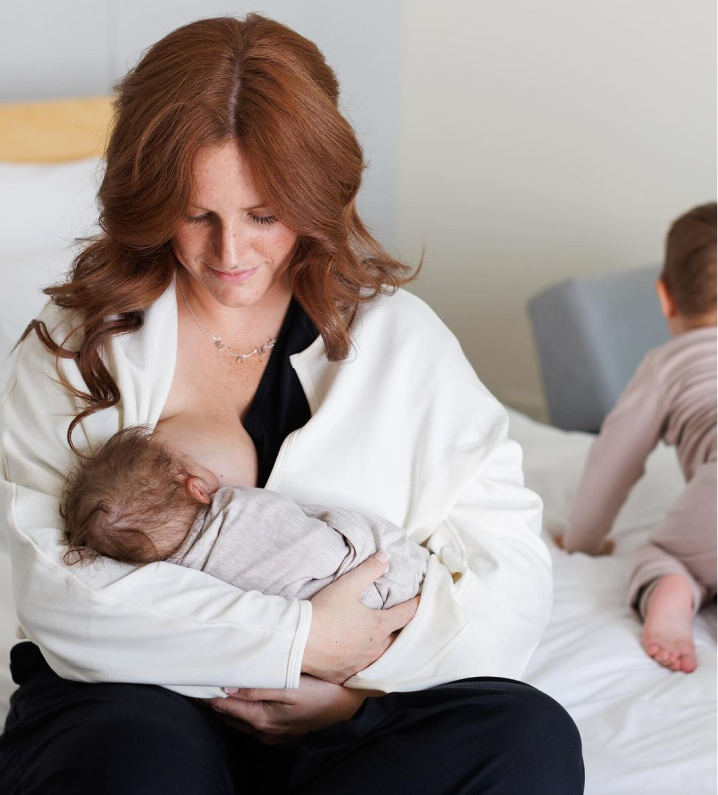 A woman breastfeeding a baby, with another child in the background on a bed.