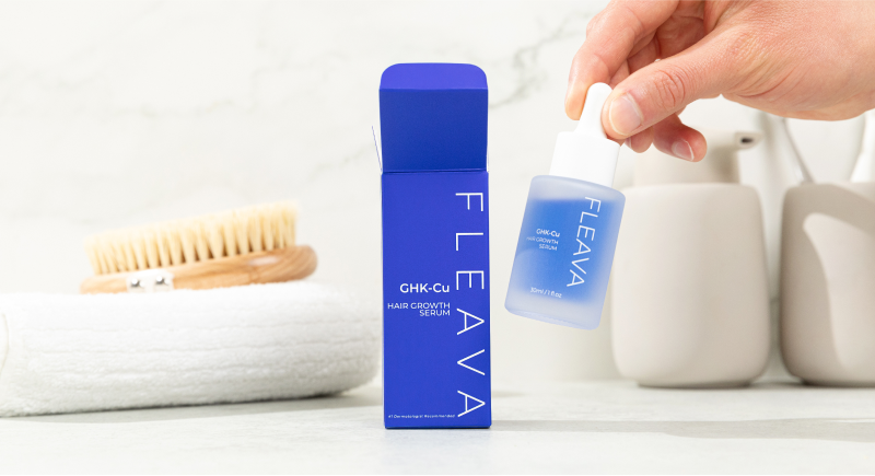 A hand holding Fleava GHK-Cu Hair Growth Serum next to its blue box within a bathroom setting.