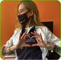 Person wearing a mask and lab coat, making a heart shape with hands.