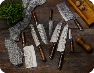 A set of patterned kitchen knives with wooden handles on a wooden counter, alongside herbs and a pepper grinder.