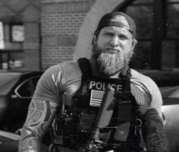 Police officer with tactical gear and tattoos standing outside in front of a building.