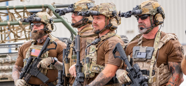 Four soldiers in tactical gear with helmets and night vision devices lined up outdoors.