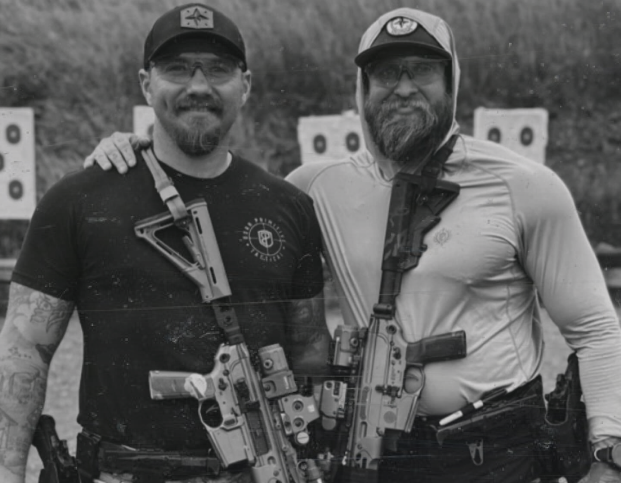 Two men with rifles at an outdoor shooting range, smiling together.