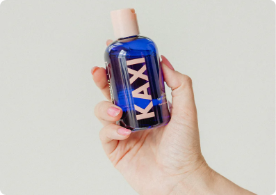 Hand holding a blue bottle labeled 'KAXI' with a light pink cap.