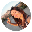 A person taking a selfie outdoors, smiling and making a peace sign, with a scenic background.