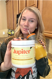 Person holding a Jupiter supplement container and orange drink in a kitchen.