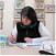 A person writing at a desk, surrounded by framed pictures.