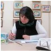 Woman seated at a table, writing on paper, surrounded by framed pictures.