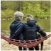 Two people sitting on a hammock by a lake in a green, wooded area.