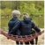 Two people with gray hair sitting together on a hammock by a lake in a wooded area.