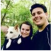 Two people and a dog smiling outdoors in a green, wooded area.