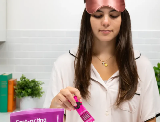 Woman in pajamas and sleep mask holding a small pink bottle in a kitchen.