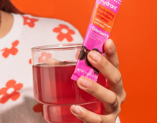 Person holding a glass with a packet of elderberry-flavored drink mix against an orange background.