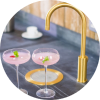 Two pink cocktails in elegant glasses beside a gold faucet on a gray countertop.