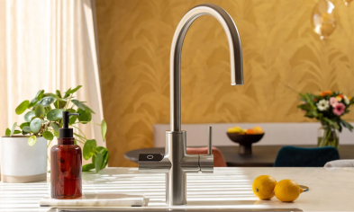 Modern kitchen faucet with a curved design, with fruits and plants in the background.