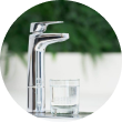 A sleek faucet next to a filled glass on a countertop with a green plant background.