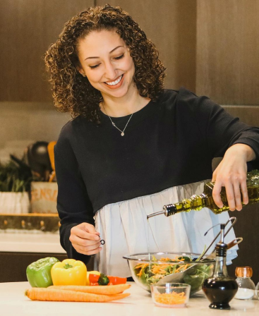 Woman smiling and pouring olive oil into a salad bowl in a kitchen setting.