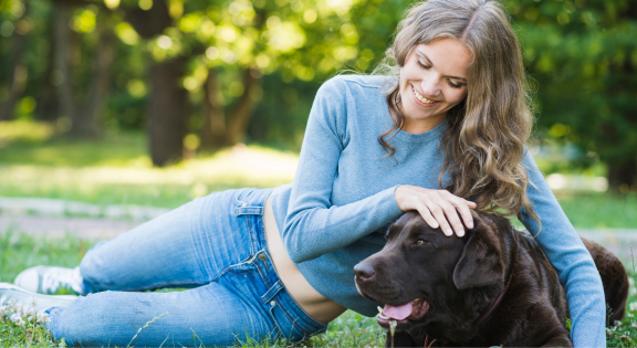 Woman laying on grass petting a brown dog in a park.