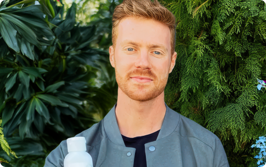 A person standing outdoors holding a white bottle, surrounded by lush green foliage.