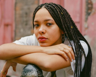 A person with braided hair leans on a railing in front of a rustic background.