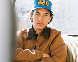 A person wearing a brown jacket and blue cap sitting indoors.