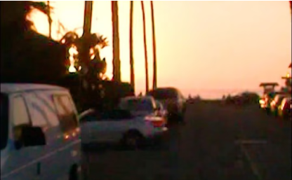 Street with cars parked at sunset, palm trees lining the background.