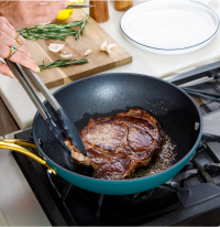 Cooking steak in a frying pan with rosemary, garlic, and lemon on a chopping board.