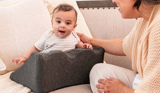 A baby sits supported by a foam cushion on a sofa, with an adult beside them.