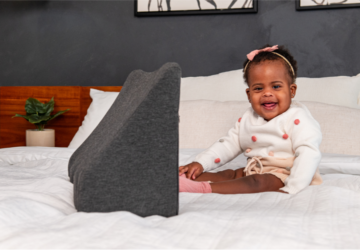 Baby in white sweater sitting on a bed, smiling beside a gray wedge pillow.