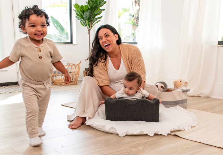 A mother smiling with two young children playing in a bright room.