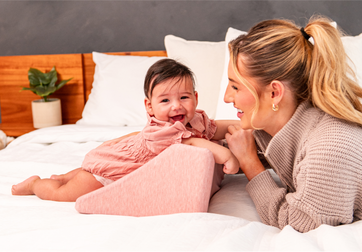 A smiling baby and woman on a bed with a pink cushion.