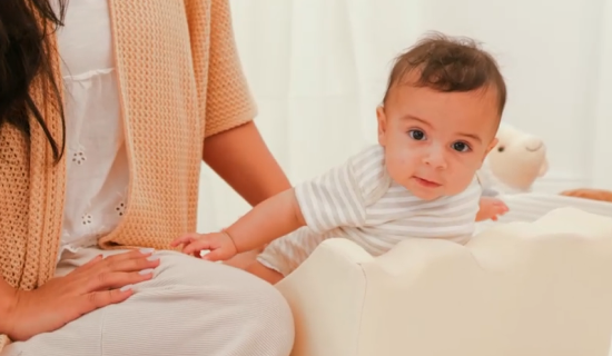 A baby in a striped onesie sitting beside a woman on a pastel bed.