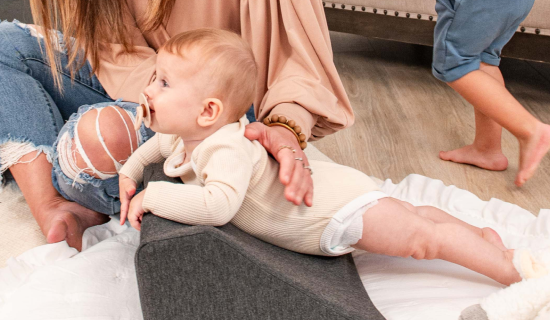 A baby lying on a wedge pillow with a pacifier, next to an adult.