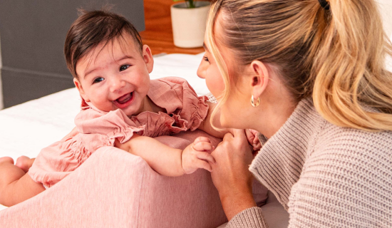 A smiling baby in a pink outfit is playing with and smiling at an adult woman.