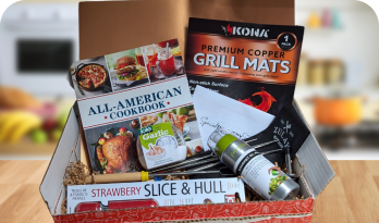 Gift box with a cookbook, grilling supplies, and kitchen gadgets on a wooden table.