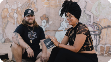 Two people sitting outdoors in front of a mural, smiling and interacting.