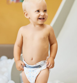 A smiling baby wearing a diaper, standing in a play area.